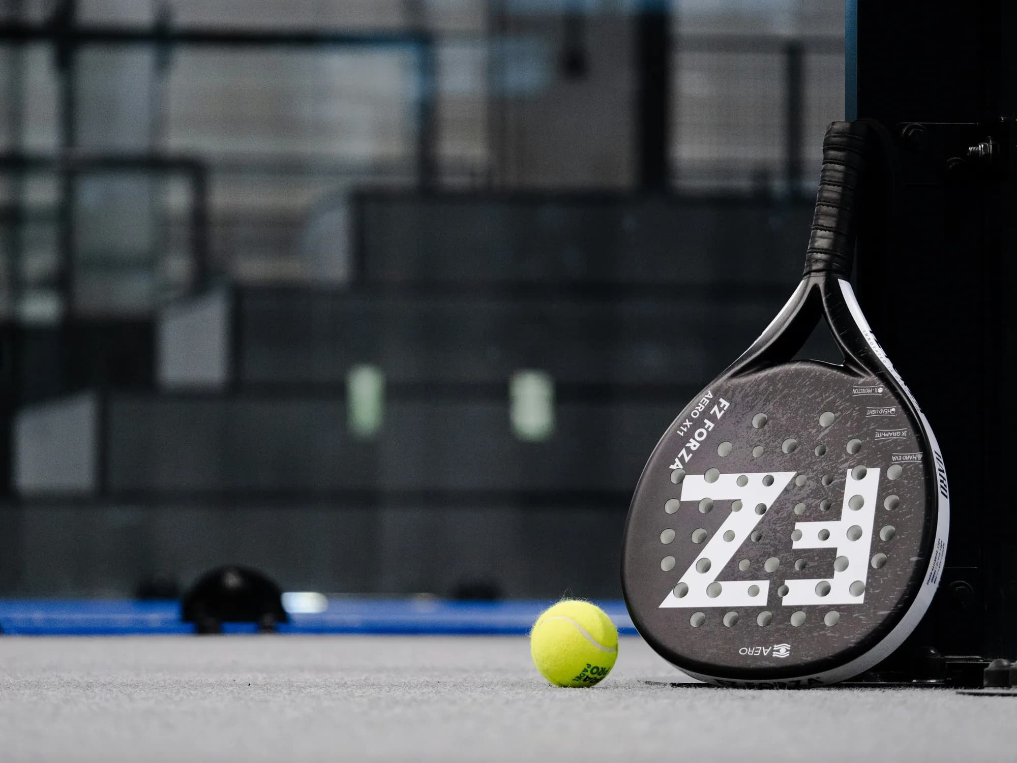 Une raquette de padel et une balle jaune au bord d’un terrain.
